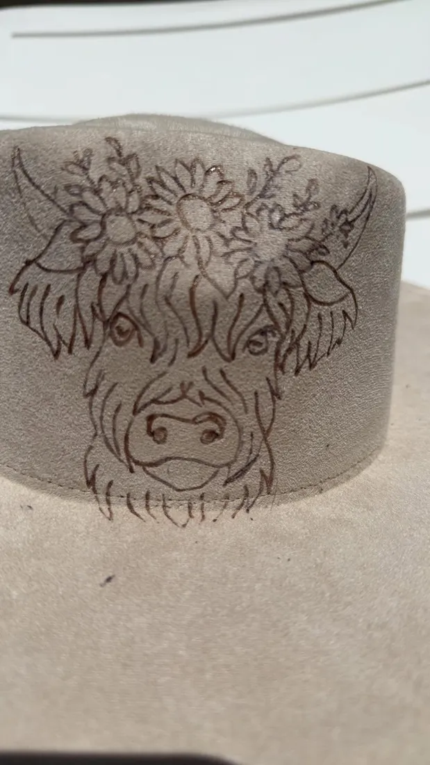 A highland cow wearing a floral crown, hand-burned onto the brim of a suede hat.
